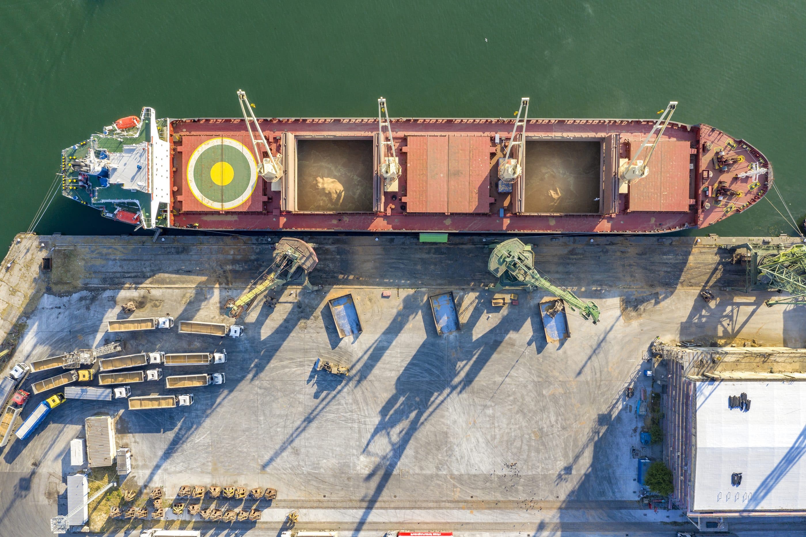 Overhead view of a bulk carrier alongside a quay, with trucks and cargo handling equipment on the dock